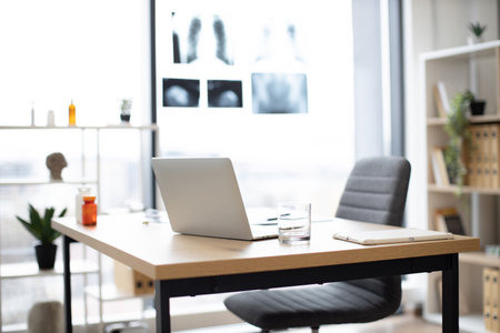 Half empty glass of water standing on writing desk between laptop and digital tablet on doctors office background. General practitioners recommending water balance maintenance for body health.の写真素材