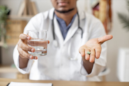 Focus on green round pill being held by multiracial male doctor sitting at writing desk with laptop on office background. Professional medical worker giving medicine and glass of water for treatment.の写真素材