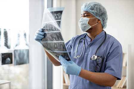 Mindful multiethnic man in protective garment holding detailed CT scan image of body part while...