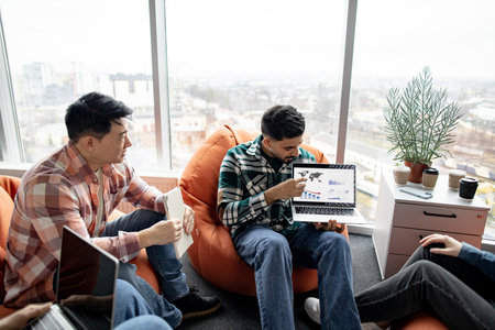 Focus on multicultural men talking about graphic materials on computer while female colleagues sitting on poufs. Business experts analyzing market growth using data visualization in office interior.の写真素材