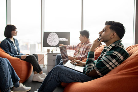 Focus on thoughtful indian male sitting on beanbag with laptop while multicultural coworkers dealing...