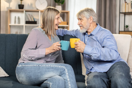 Portrait of happy senior family of two in casual attire sitting on comfortable sofa and smiling at...