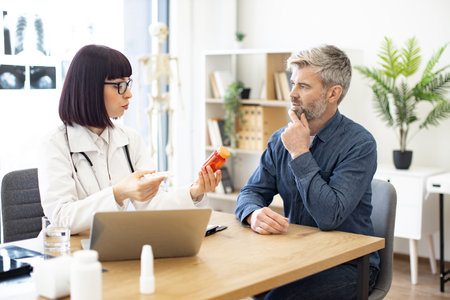 Female doctor providing expert check-up and giving medicine to middle-aged patient in hospital office. Focused man listening attentively to recommendations regarding medical history kept on laptop.の写真素材