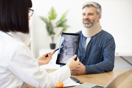 Back view of female doctor holding digital tablet while sitting in front of smiling caucasian man in cervical collar. Health professional consulting mature patient about neck injury using CT scan.の写真素材