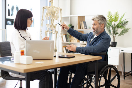 Brunette female in lab coat talking to bearded male in wheelchair while sitting at desk in consulting room of hospital. General practitioner giving medical advice about treatment using skeleton.の写真素材