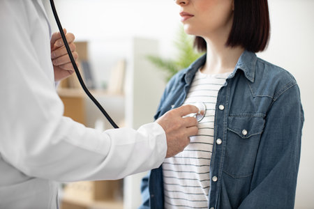 Cropped view of young brunette in denim jacket attending medical appointment at doctors office in health center. Male physician examining female patient with stethoscope in consulting room interior.の写真素材