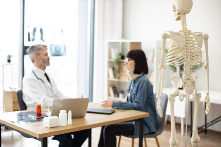 Focus on back view of teaching model located in consulting room of private practice with adults talking in background. Experts using skeleton as representation of human anatomy for patient education.の写真素材