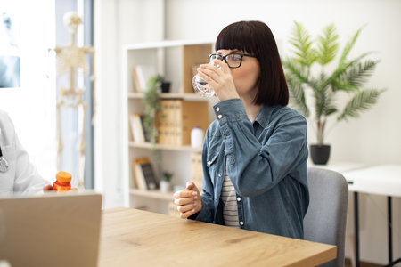 Pretty caucasian female in casual wear drinking water while seating at writing desk in doctors office of health clinic. Beautiful woman taking oral medication that being swallowed with liquid.の写真素材