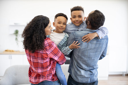 Back view of multicultural spouses holding preschool boys in arms while having break from playing board games. Friendly family planning after-dinner walk before leaving cozy home on sunny day.の写真素材