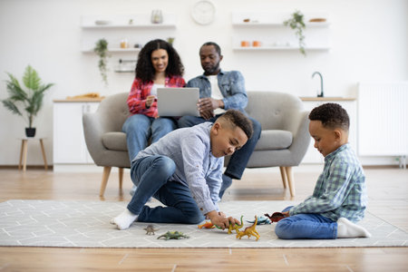 Focus on two multiracial brothers sitting on carpet in living room while parents using laptop on sofa in home interior. Cheerful kids playing with toy dinosaurs while adults watching online TV show.の写真素材