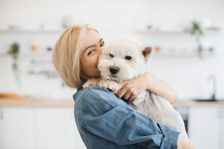 Portrait of smiling blonde lady with West Highland White Terrier in arms posing on background of spacious kitchen. Happy caucasian pet owner rewarding good behavior during day with gentle hug.の写真素材