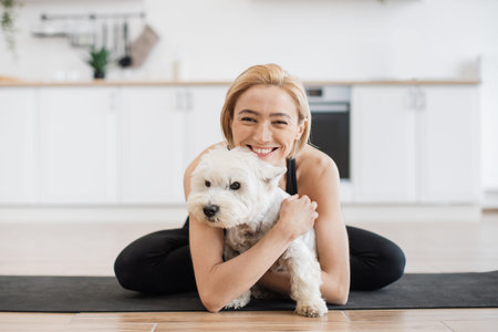 Affectionate adult female in yoga activewear embracing white terrier while sitting cross-legged on rubber mat indoors. Healthy lady awarding sweet pet with hug for good behaviour during home workout.の写真素材