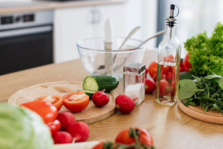 Close up view of assorted raw vegetables and leafy lettuces left on cutting board near bowl with cutlery on kitchen table. Natural ingredients for homemade salad with classic dressing ready for use.の写真素材