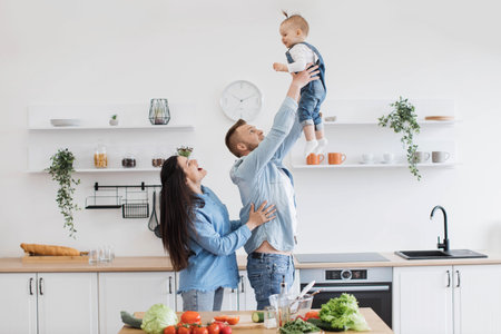 Handsome strong father raising child high in air while excited mother cheering her daughter up in dining room. Emotional couple and baby girl increasing happiness and fun during cooking meal at home.の写真素材