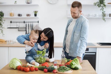 Focused infant girl holding metal spoons and trying to mix salad while beautiful mother teaching how to do it. Proud mother and father encouraging little baby learning new cooking skills in kitchen.の写真素材