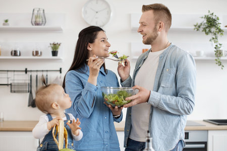 Positive husband and wife in everyday clothes feeding each other with fresh vegetable salad in stylish white kitchen while cute baby watching at them with curiosity. Concept of food and family unity.の写真素材