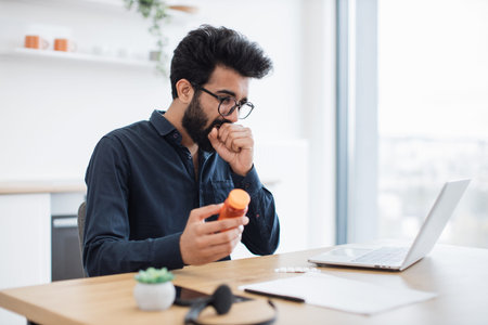 Coughing indian adult in glasses staring at wireless computer screen while spending intent hours of...