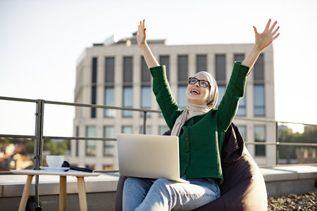 Close up view of muslim lady in traditional clothing holding fist high in air while scanning laptop screen on roof terrace. Excited young person in glasses expressing joy after reading email outdoors.の写真素材