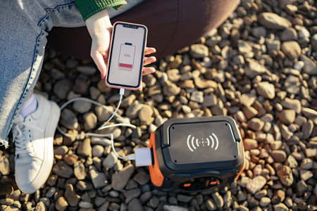 Top view of mobile device in hand connected to portable power station via white cord outdoors....
