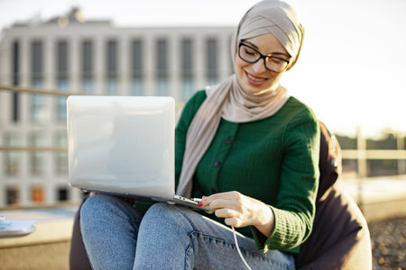 Close up view of smiling arabian woman in hijab holding laptop on knees while putting charge cable into port on terrace. Cheerful young lady powering portable device with off-grid electricity.の写真素材