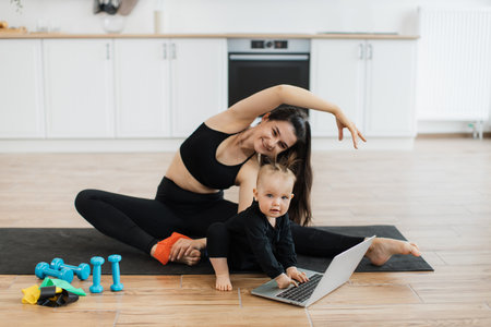 Portrait of cheerful woman and child posing in yoga outfits while doing stretching exercises on mat in modern kitchen. Active mommy improving flexibility while curious daughter studying computer.の写真素材