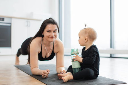 Close up view of strong healthy woman in plank pose smiling at infant girl on yoga mat playing with...
