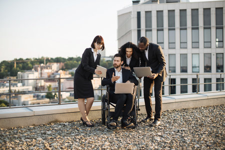 Multiracial global experts cooperating on panoramic terrace of office building and agreeing plan regarding new construction. Caucasian man with disability looking at digital tablet and gesturing hand.の写真素材
