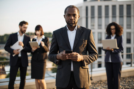 Portrait of successful african american entrepreneur holding mobile phone in hands and smiling at...
