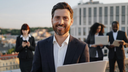 Portrait of cheerful caucasian businessman looking at camera with laptop in hands and smiling. Diverse group of specialists dressed in suits using modern devices for creating project draft drawing.の写真素材