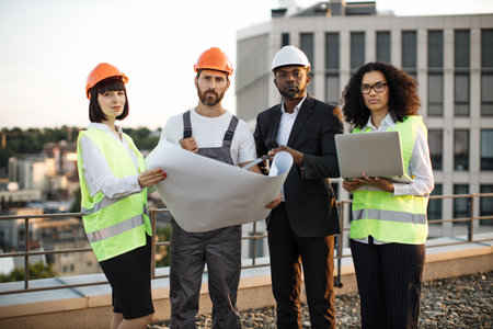Portrait of multicultural group of developers posing on camera with portable gadget and blueprints. Four international colleagues in vests and hardhats smiling while meeting at panoramic terrace.の写真素材