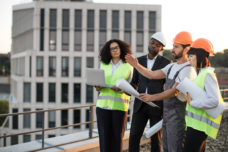 Four multiracial engineers with technical drawings and laptop discussing place for construction of new multi-apartment complex. Workman talking and pointing with hand while others listening carefully.の写真素材