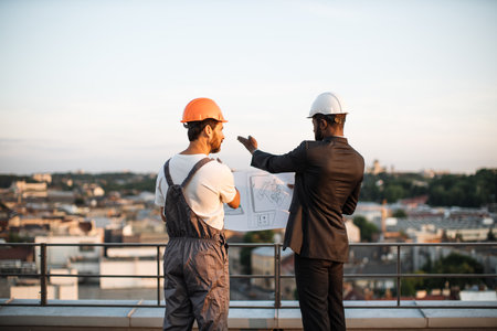 Back view of foreman in grey uniform and business investor in black suit watching together at engineering drawings while standing on panoramic terrace. Male builders discussing real estate project.の写真素材