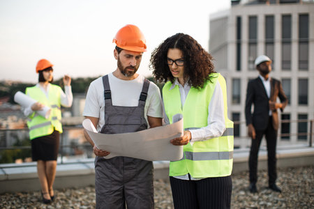 Bearded builder in hardhat presenting technical blueprints to african american supervisor while two...