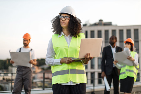 African american female architect in vest and hardhat standing on rooftop with laptop in hands and looking aside. Multiracial builders with blueprints watching building process at construction site.の写真素材