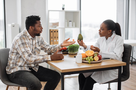 Handsome multiracial man in casual clothes visiting female dietitian in doctors office of medical center. Curious patient asking about avocado usage for special dietary requirements on consultation.の写真素材