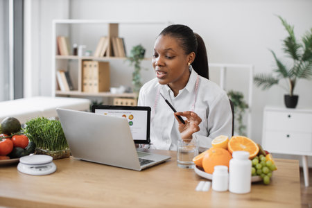 Focused multicultural expert with meal plan on tablet conducting web meeting using portable laptop at work. Skilful female nutritionist explaining details of slimming program for client online.の写真素材