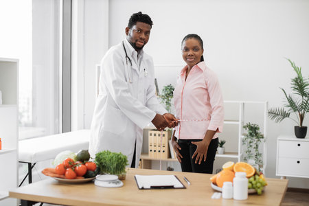 Full length view of multiracial specialist taking measurements of ladys waist at dietitians office in clinic. Attentive male professional evaluating clients health by estimating body composition.の写真素材