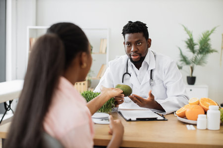 Focused african american man in white coat holding avocado while talking with female client in consulting room. Medical specialist in food encouraging to enriching eating with fruit full of nutrients.の写真素材
