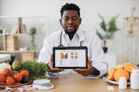 Confident african american male in doctors coat showing tablet with diet plan on screen while working at hospital. Positive dietitian planning nutrition program on clients personal preferences.の写真素材
