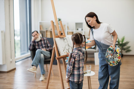 Attractive woman introducing little girl to fine arts while showing sketch of senior man on canvas at home. Caring young mother helping cute daughter with color choice for grandpas portrait.の写真素材