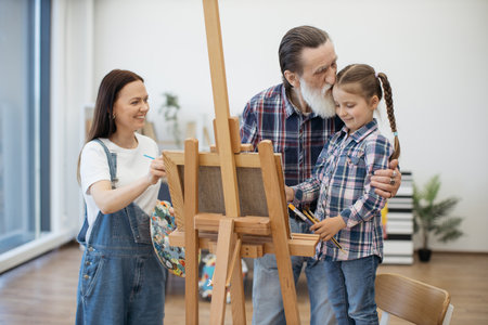 Loving aged grandfather in casual wear kissing little kid while smiling lady doing sketches on cloth in room. Affectionate grown-ups and child strengthening family bonds while engaging in arts.の写真素材