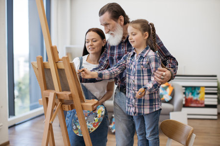 Confident aged gentleman pointing at canvas placed on easel while little girl asking about colors of art project. Curious mother and daughter peering at sketch of human made by granddad indoors.の写真素材
