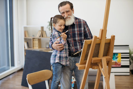 Caring grandparent cuddling little child with paintbrush while doing easel-type portrait in home art studio. Grey-haired mature man sharing creative skills with active granddaughter on weekend.の写真素材