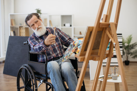 Grey-bearded retired man with disability sitting in front of wooden easel and examining drawing. Aged man spending pastime alone painting on canvas in bright spacious living room.の写真素材