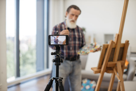 Blurred senior blogger broadcasting while painting new beautiful picture on easel at cozy art studio. Imposing elder male dressed in jeans and checkered shirt communicating with followers on internet.の写真素材