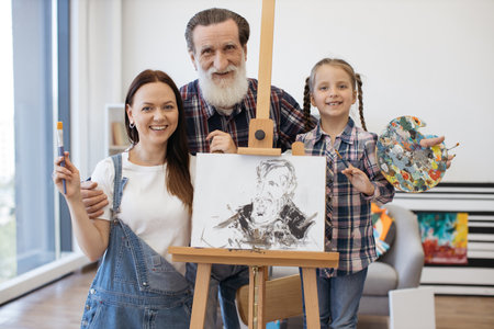 Portrait of loving family of mother, granddad and granddaughter looking together at camera and smiling. Kind man of mature age holding colored palette while mother with girl posing with paintbrushes.の写真素材