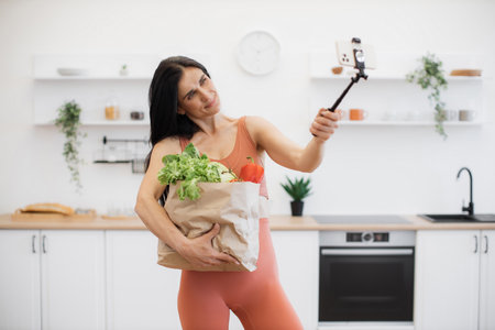 Cheerful woman in activewear posing at camera of mobile on monopod while holding carrier bag with food. Sports blogger with fresh vegetables and greens for dinner shooting photos after home workout.の写真素材