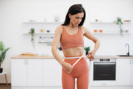 Attractive woman in good health measuring waist with tape after working out at home in yoga activewear. Athletic person steering in direction of improving well-being via food habits and fitness.の写真素材