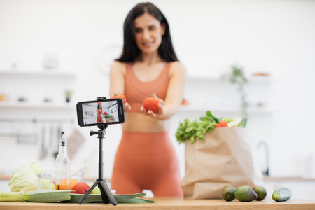 Focus on smart device with working camera app fixed by tripod on kitchen table with slender female in background. Happy vlogger with tomatoes shooting video content about sports nutritional foods.の写真素材