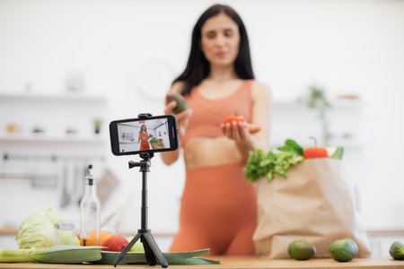 Selective focus of cell phone on tripod being used by cheerful lady with fresh salad ingredients from shopping bag. Fitness and nutrition blogger sharing tips on choosing proper products for meals.の写真素材
